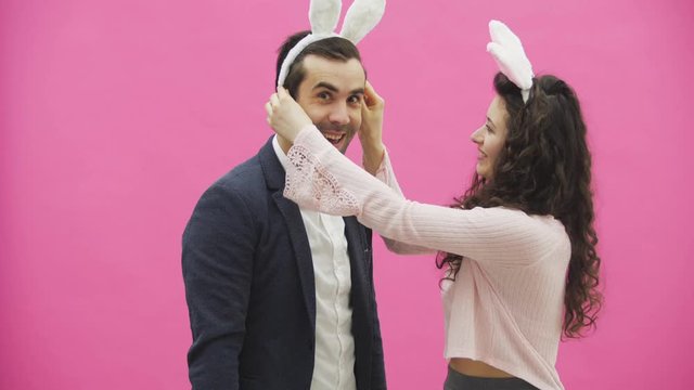 Young lovers couple on the pink background. With hackneyed ears on the head. During this, the girl wears rabbit ears to her husband. After that, they begin to make a photo of sephi on the phone. Very