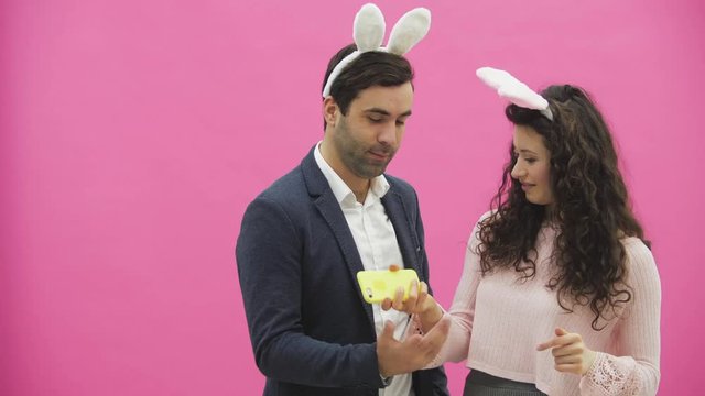 Young lovers couple on the pink background. With hackneyed ears on the head. During this, they make a photo of sephi on the phone showing various rabbit movements and rejoice.