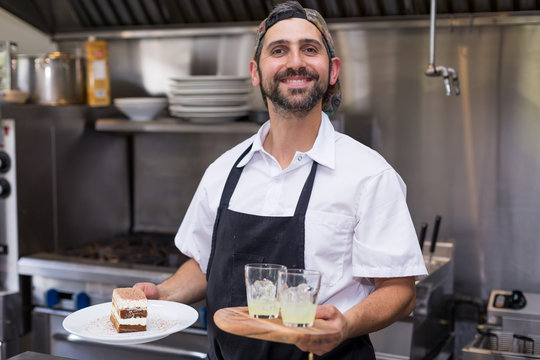A Happy Smiling Male Chef Holding Tiramisu And Limoncello In A Restaurant Kitchen.
