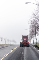 Fototapeta na wymiar Dark red big rig semi truck tractor moving ahead on foggy winter road with trees on the sides