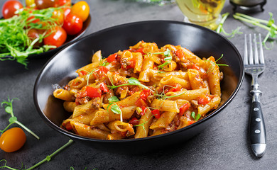 Penne pasta in tomato sauce with meat, tomatoes decorated with pea sprouts on a dark table.