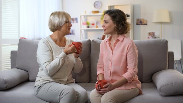 Mother and daughter talking at home, relaxing on sofa with cups of coffee, trust