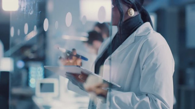  In the Research Laboratory Smart and Beautiful African American Female Scientist Wearing White Coat and Protective Glasses Writes Formula on Glass Whiteboard, References Her Tablet Computer