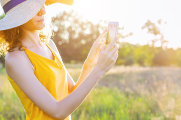 Happy smiling woman female in summer hat and lite yellow summer dress doing picture photo of nature by smartphone, mobile phone in green park outdoor. Summer, spring active outdoor leisure concept.