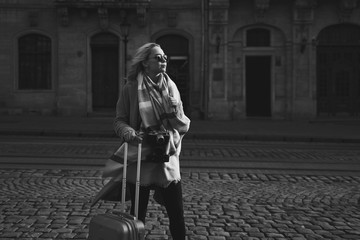 Young beautiful blond woman tourist with a wheeled travel bag and vintage film camera standing on Rynok square in Lviv, Ukraine