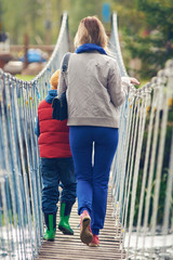 Young European woman and her son are walking in the picturesque park. They are going through the rope bridge across the raging river.