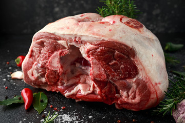 Raw lamb leg on black stone table with herbs. garlic, rosemary, chilli, salt, pepper, Bay leaf.