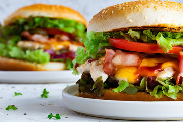 Fresh tasty chicken burger on white table. Two chickenburgers with bacon, lettuce, tomato, sauce, cheese, close-up
