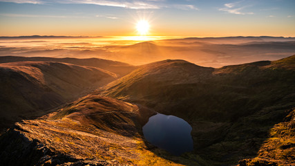 A stunning orange yellow sunrise from Sharp Edge on Blencathra, Lake District, Cumbria with reflection in lake tarn and cloud inversion over mountains in the distance