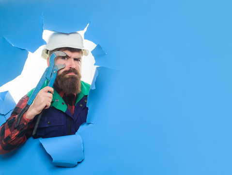 Bearded man with tools in hand. Through paper. Plumbing and renovation. Tools or repair service. Worker with pipe wrench. Bearded man in helmet. Uniform. Bearded man through paper.
