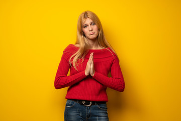 Blonde woman over yellow wall keeps palm together. Person asks for something