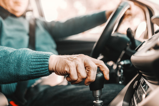 Close up of senior man holding one hand on gearshift and other on steering wheel while sitting in his car. Selective focus on hand.