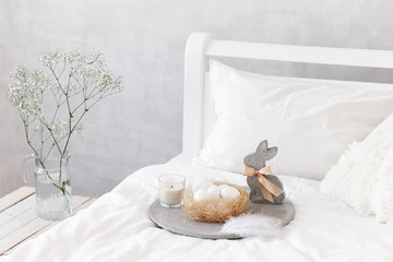 White modern bedroom with Easter decoration. Bed with white bedding set, concrete tray, nest with white eggs, decorative bunny figure, candle and  gypsophila flowers in a glass vase.