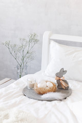 Fototapeta na wymiar White modern bedroom with Easter decoration. Bed with white bedding set, concrete tray, nest with white eggs, decorative bunny figure, candle and gypsophila flowers in a glass vase.