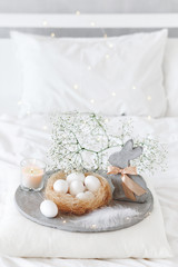 Fototapeta na wymiar White modern bedroom with Easter decoration. Bed with white bedding set, pillows, concrete tray, nest with white eggs, decorative bunny figure, candle and gypsophila flowers.