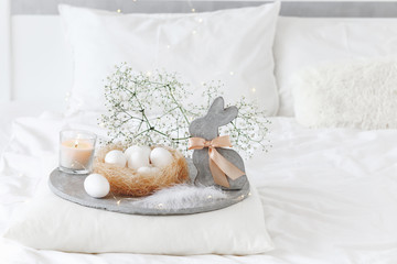White modern bedroom with Easter decoration. Bed with white bedding set, pillows, concrete tray, nest with white eggs, decorative bunny figure, candle and  gypsophila flowers.