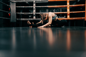 Full length of Caucasian female boxer girl in sportswear stretching on floor in ring.