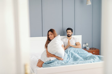 Obraz premium Angry woman sitting in the bed and holding pillow with back turned to her husband who begging her for forgiveness. Bedroom interior.