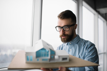 Young pensive architect holding a home model in loft modern office. Architectural project concept. University student planning engineering design, looking for solution, brainstorming at workplace