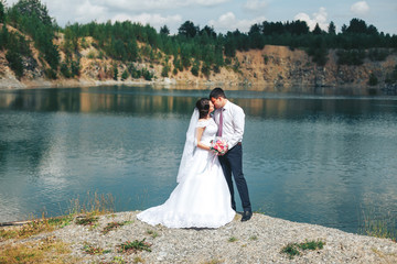 Fototapeta na wymiar A beautiful newlywed couple standing near a blue clear lake. Tender and touching photo of the bride and groom