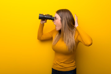 Young woman on yellow background and looking in the distance with binoculars