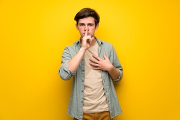 Teenager man over yellow wall showing a sign of silence gesture putting finger in mouth