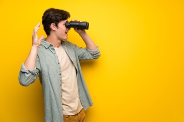 Teenager man over yellow wall and looking in the distance with binoculars