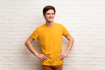 Teenager man over white brick wall posing with arms at hip and smiling