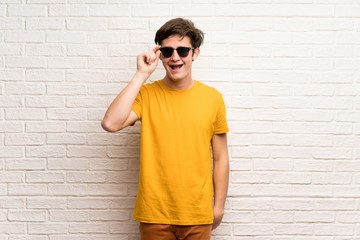 Teenager man over white brick wall with glasses and surprised