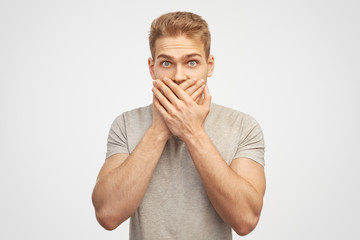 Incredulous attractive man covers mouth, doesnt want to spread rumours, shocked to hear sudden news from interlocutor, dressed in casual tshift isolated on white wall - obrazy, fototapety, plakaty