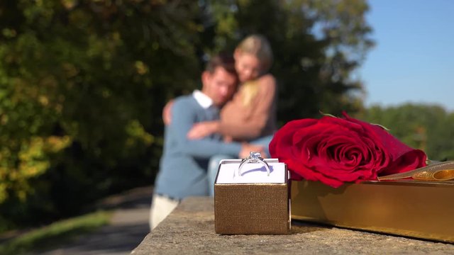 Closeup on a wedding ring, a present and a red rose in a park - a happy couple hugs in the blurry background
