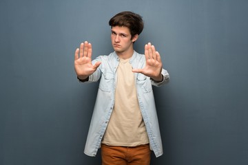 Teenager man with jean jacket over grey wall making stop gesture and disappointed