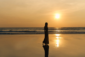 Contrast silhouette of a young slender woman against the background of a Sunny sunset, the sea and the sandy beach. Warm evening tones and reflections of the figure on the wet sand