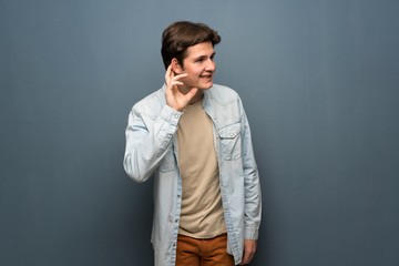 Fototapeta premium Teenager man with jean jacket over grey wall listening to something by putting hand on the ear