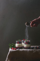 Homemade classic dessert Panna cotta with raspberry and blueberry berries and jelly in jars, decorated by mint with sprinkling sugar powder over linen table cloth with grey wall at background.