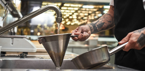 Cooling down. Close up photo of chef's hands with several tattoos holding cooked pasta in a colander under water over sink in the kitchen