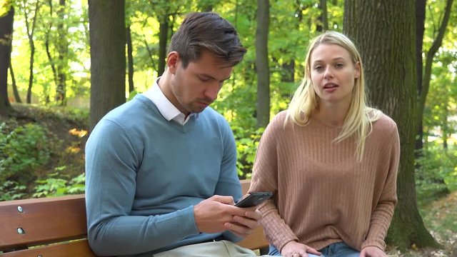 A man and a woman sit on a bench in a park, the man is absorbed with his smartphone, the woman is angry at him