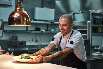 Ready to serve. Young smiling chef with tattoos on his arms, in black apron holding ready dish in...