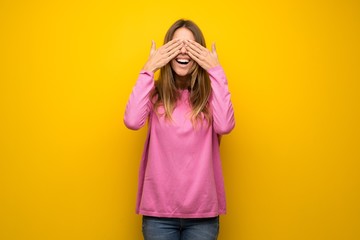 Woman with pink sweater over yellow wall covering eyes by hands. Surprised to see what is ahead