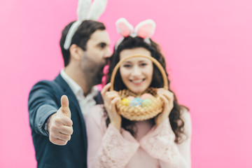 Young couple on pink background. Smiling looking at the camera. On the head, the hare of the ears. The spring holds a basket of eggs. A man shows a close-up class. Easter.