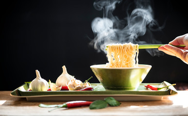 Hand uses green chopsticks to pickup noodles with steam and smoke in bowl on black background,...
