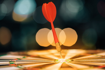 Red dart arrow hitting in the target center of dartboard with city light bokeh background. Target business, achieve and victory concept.