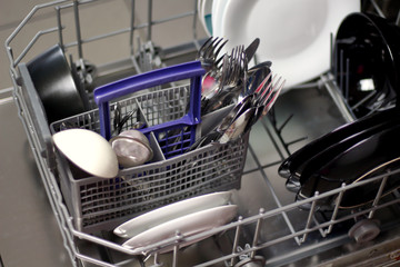 Dishwasher with white and black plates with bowls, forks, spoons, knives in a stand basket, clean washed dishes in a dishwasher