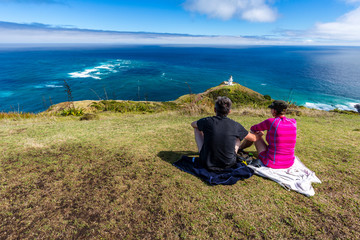 Fototapeta premium Couple enjoying the view Lighthouse at Cape Reinga, with Tasman Sea and Pacific, Northland, New Zealand