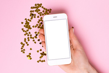 Mobile phone with white empty screen in female hand on pink color paper background with golden confetti