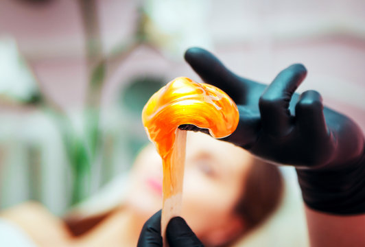 Close-up hands of cosmetologist in black gloves preparing golden colored waxing paste on spatula for sugaring depilation, hair removal beauty procedure.