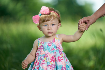 Cute little girl in dress adorned with flowers holds the hand of adult and looks into the camera,...