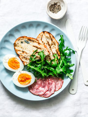 Delicious breakfast or snack - salami sausage, boiled egg, arugula, grilled bread and coffee on a light background, top view