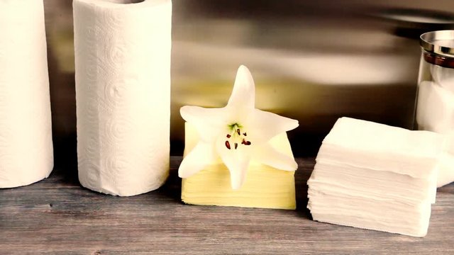 Piece of interior with paper disposable products - paper towel, napkins, tissue, cotton pads and a flower of lily on wooden table against steel background with reflections. Slow pan.
