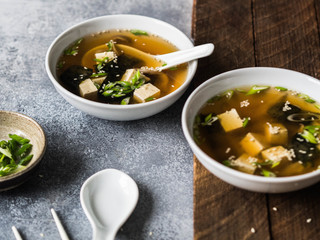Japanese miso soup with oyster mushrooms in a white bowls with a spoon and white chopsticks on a grey and wood backgrounds.
